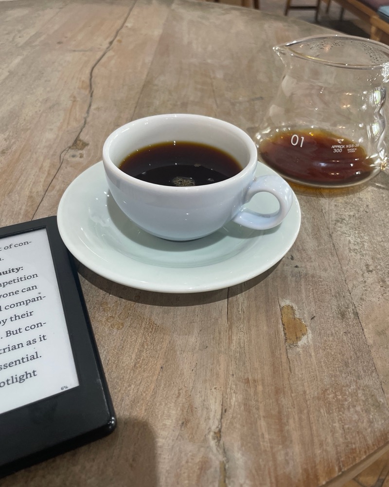 A cup of filter coffee, a small carafe, and an e-reader on a wooden table